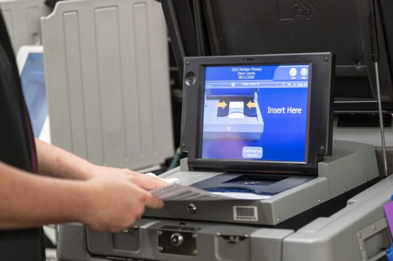 A person holds a paper ballot at a voting machine. Its touchscreen displays instructions for the 2026 partisan primary in Dane County, including "Insert Here."